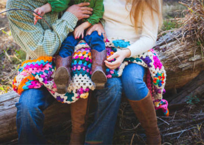 A family sits on a log as the father holds a young child wearing boots on his lap and his mother rests her hand on the child