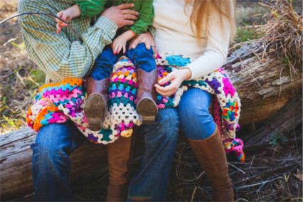 A family sits on a log as the father holds a young child wearing boots on his lap and his mother rests her hand on the child