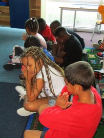 Seven children sit on the floor praying with their hands folded
